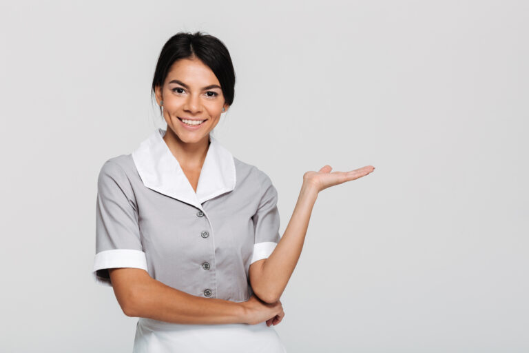 Close-up portrait of young happy housekeeper in uniform showing empty palm while looking at camera, isolated on white background