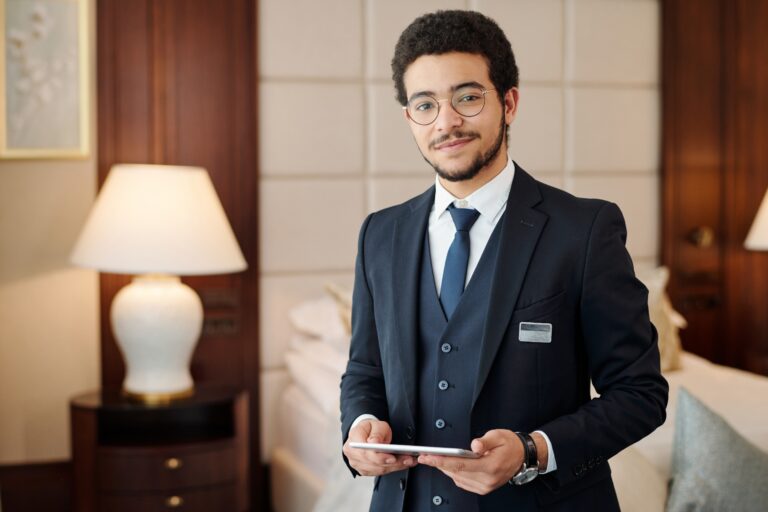 Young confident head manager of luxurious hotel looking at camera while standing against lamp and comfortable double bed in room