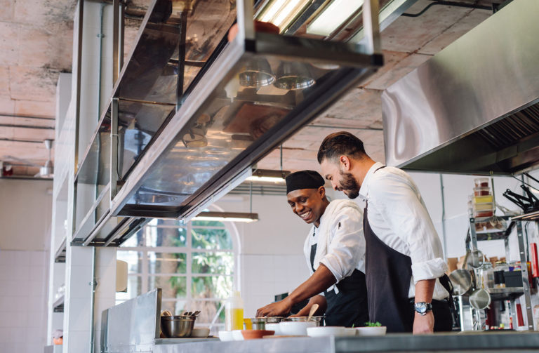 Happy male chefs cooking food at cafe kitchen and talking. Two cooks preparing food in restaurant kitchen.