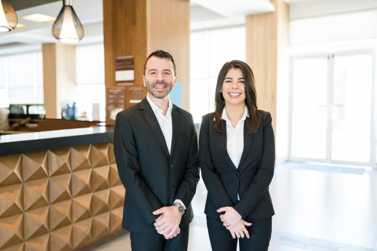 Confident attractive male and female managers smiling while making eye contact against reception desk in hotel